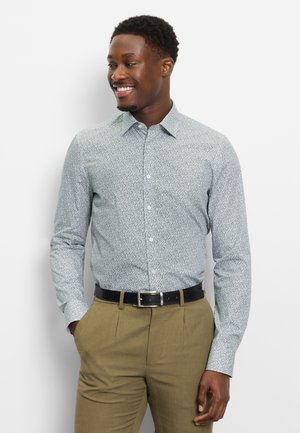 Uomo sorridente con le mani in tasca, indossa una camicia chiara a fantasia con bottoni, pantaloni kaki e una cintura nera, in piedi su sfondo bianco.