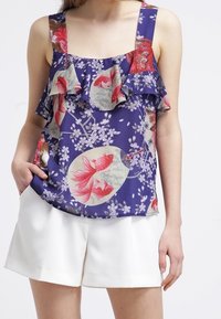 Kvinna klädd i en lila kortärmad topp med rosa fiskar och blommönster, tillsammans med vita shorts framför en enfärgad bakgrund.