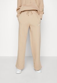 Pantalones de chándal beige hechos de una tela suave, con cintura ajustable, piernas anchas y un pequeño logo blanco de Nike en la pierna izquierda.