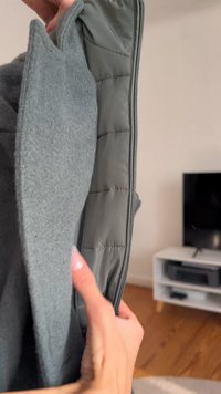 Γκρι σακάκι με μαλακή εξωτερική επιφάνεια από μάλλινο ύφασμα και επένδυση από πλισέ, που διαθέτει φερμουάρ και λεία υφή, κρατημένο ανοιχτό από ένα χέρι.