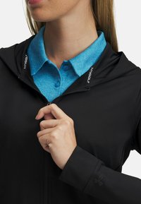 Black zippered jacket with a hood; features a blue collared shirt underneath. Textured fabric and logo detail visible on the sleeve.