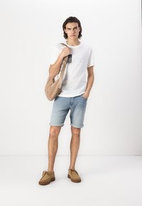T-shirt en coton blanc avec impression graphique, short en denim bleu clair, chaussures en suede marron et sac à bandoulière beige. Design simple, coupe décontractée.