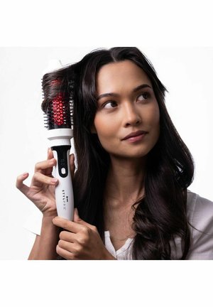 Woman with long dark hair using a white heated round brush labeled "STYLPRO" to style a section of her hair against a white background.