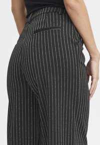 Črne hlače s pinstripi, gladke teksture, visokega pasu, s stranskimi žepi in krojem po meri, prikazane s stranske strani.