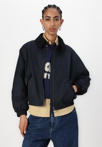Navy bomber jacket with a corduroy collar, zip front, and ribbed cuffs, layered over a beaded sweater and beige shirt. Denim pants.