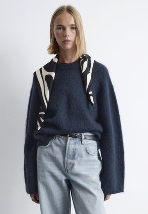 Dunkelblauer, flauschiger Pullover mit weiten Ärmeln, kombiniert mit hellblauen Jeans. Ein schwarz-weiß gemusterter Schal hängt über der Schulter.