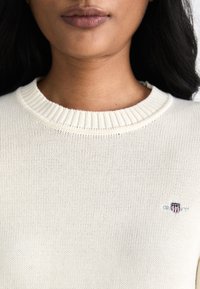 Kreemivärvine kudumipullover ribbiga kaelusega ja väikese brodüüritud logoga rinnal. Suurendatud vaade kangastektsioonile.