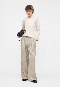 Άτομο που φοράει μπεζ μπλούζα με μακριά μανίκια και παντελόνι με φαρδιά πόδια, κρατώντας μια μαύρη τσάντα με τρουκς, στέκεται μπροστά σε απλό ανοιχτό φόντο.