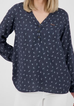 Kobieta nosząca granatową bluzkę z długim rękawem z małymi białymi wzorami ptaków i trzema czarnymi guzikami na dekolcie w kształcie litery V, w połączeniu z białymi spodniami.