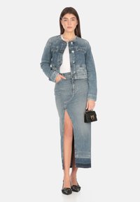 Giacca di jeans con chiusura a bottoni, colore azzurro chiaro, due tasche sul petto, abbinata a una lunga gonna di jeans con uno spacco laterale e una borsa nera.