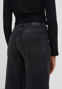 Jean en denim noir à taille haute avec une coupe large. Présente une texture lisse, deux poches arrière et une étiquette de ceinture en cuir noir.