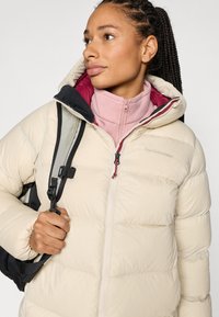 Beige Steppjacke mit Kapuze und gestepptem Design, mit einem kontrastierenden weinroten Futter, getragen über einem hellrosa Reißverschlussoberteil.
