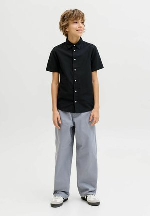 Rövid, göndör hajú fiú fekete, rövid ujjú, gombos ingben, laza szürke nadrágban és fehér sportcipőben áll, mosolyogva.