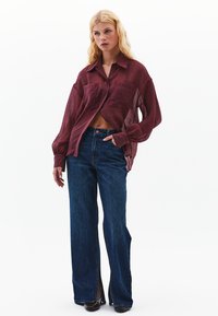 Burgundy, genomskinlig skjorta med knappar och två frampocket, kombinerad med högmidjade, vida, mörka denimjeans med sidoklappar.