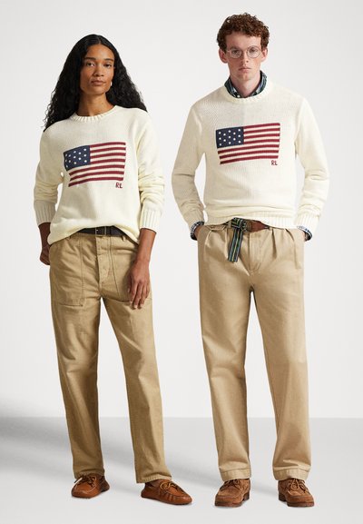 Μπεζ πουλόβερ με κεντημένες σημαίες των ΗΠΑ, σε συνδυασμό με μπεζ παντελόνια. Το αρσενικό μοντέλο φοράει πουκάμισο με σχέδιο κάτω από το πουλόβερ. Καφέ παπούτσια.