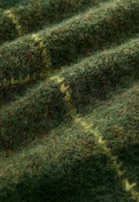 Wollstoff mit einer strukturierten Oberfläche in dunklen Grün- und Brauntönen, mit einem auffälligen gelblichen Muster, das entlang der Fasern verläuft.