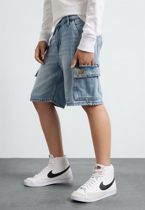 Osoba nosząca jasnoniebieskie denimowe szorty cargo i białe wysokie trampki Nike, stojąca z jedną ręką w kieszeni.