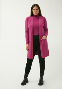 Heller rosa langer Cardigan mit offenem Vorderteil, gerippten Bündchen und zwei Vorderaschen, kombiniert mit einem rosa Rollkragenpullover sowie schwarzen Hosen und Stiefeln.
