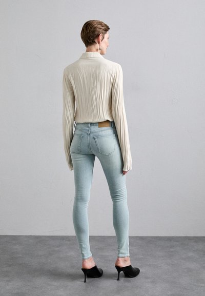 Beige plisserte langermet topp kombinert med lyseblå skinny jeans; modellen har på seg svarte åpne hæler, som fremhever en kroppsnær silhuett.