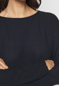 Maglione a maglia blu navy con scollatura ampia e maniche lunghe con texture a costine. Polsini arrotolati visibili sulle maniche.