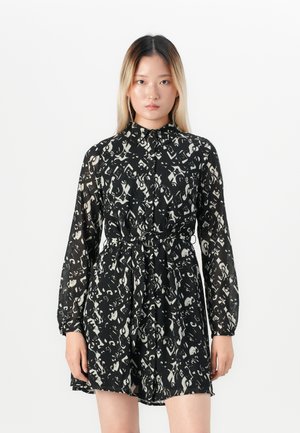 Femme avec des cheveux raides portant une robe à manches longues noire et blanche à motifs, avec un col et une ceinture, se tenant devant un fond blanc uni.