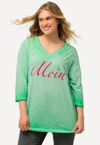 Grünes, übergroßes V-Ausschnitt-Shirt mit Dreiviertelärmeln, das das Wort "Moin" in fetter rosa Kursivschrift zeigt. Weicher, strukturierter Stoff mit Farbverlauf.