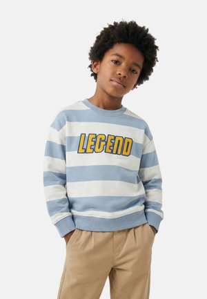 Garçon aux cheveux bouclés portant un sweat-shirt à rayures bleues et blanches avec le mot « LÉGENDE » en lettres jaunes, et un pantalon beige les mains dans les poches.