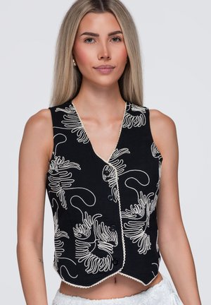 Jeune femme aux cheveux blonds et lisses portant un gilet noir sans manches avec une broderie florale abstraite blanche et des boutons noirs.