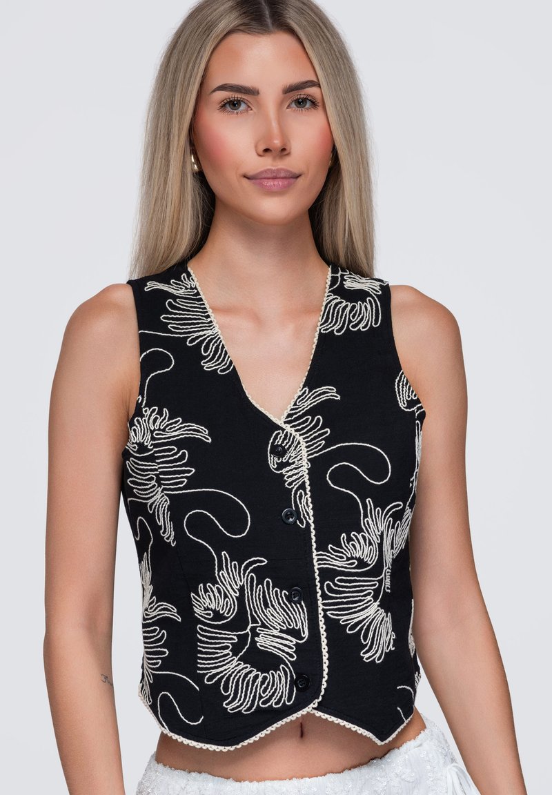 Jeune femme aux cheveux blonds et lisses portant un gilet noir sans manches avec une broderie florale abstraite blanche et des boutons noirs.