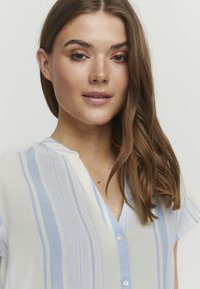 Camicia leggera con bottoni, di colore bianco con strisce verticali blu, dotata di scollatura a V e maniche corte. Tessuto morbido e trasparente.