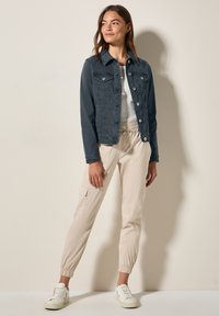 Mörkgrå denimjacka med två bröstfickor, figursydd design och vita knappar. Matcher med beige joggerbyxor och vita sneakers.