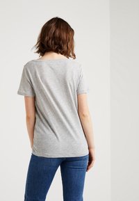 Mujer con pelo corto castaño, vistiendo una camiseta gris claro sencilla y jeans azules, de espaldas contra un fondo blanco.
