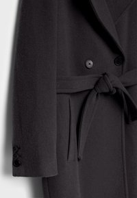 Manteau en mélange de laine noire avec col cranté, fermeture à double boutons, ceinture à nouer et poignets boutonnés. Texture douce avec une forme structurée.