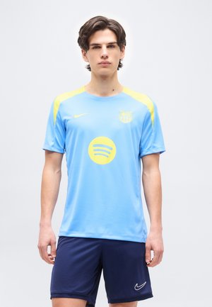 Fiatal férfi áll, világoskék és sárga FC Barcelona labdarúgó mezben és sötétkék Nike rövidnadrágban, egyszínű háttér előtt.