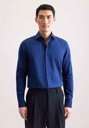 Camicia da uomo a maniche lunghe blu con colletto strutturato, trama sottile e dettagli argentati sui bottoni, abbinata a pantaloni scuri su misura.