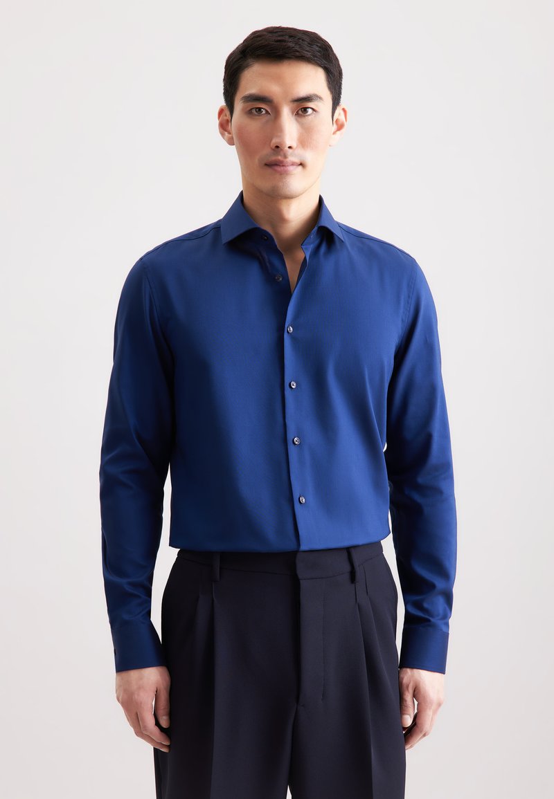 Blauw overhemd met lange mouwen en een gestructureerde kraag, subtiele textuur en zilveren knoopaccenten, gecombineerd met donkere, op maat gemaakte broeken.
