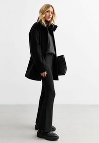 Manteau en mélange de laine noir avec un col haut, associé à un pull gris foncé et un pantalon noir évasé. Présente des bottes noires volumineuses et un sac texturé.