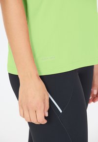 Limegrön snabb-torkande t-shirt med en slät textur. Bärs med svarta leggings som har en vit accentrand på sidan.