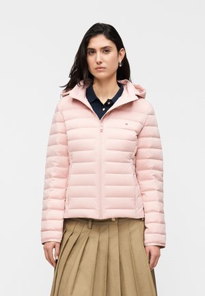 Naine kannab heledat roosat tikitud kapuutsiga jopet tumesinise särgi ja beežide plisseeritud seeliku peal, seistes ühevärvilise valge tausta ees.