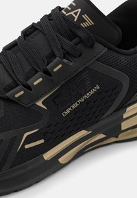 Svart sneaker med en texturerad ovandel och mesh-paneler, guldaccenter och logotyp. Tjock svart sula med kontrasterande guld detaljer.