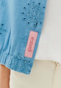 Světle modrý pletený top s vyšívanými květinovými vzory, elastickým lemem a růžovou visačkou s nápisem "GIRLitude" na levé straně.