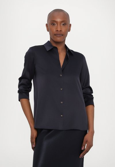 Woman with short hair wearing a dark navy button-up shirt with sleeves rolled to elbows and matching skirt, standing against a plain background.