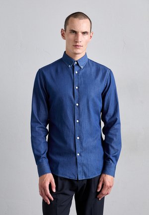 Chemise en denim bleu foncé avec un col button-down, des manches longues et des boutons blancs. Texture lisse et design ajusté.