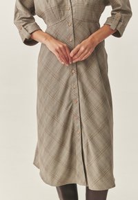 Vestito con bottoni sul davanti e maniche lunghe, in motivo a quadri beige e marrone. Presenta un corpetto aderente, una gonna plissettata e bottoni in legno.