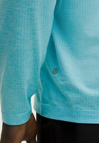 Pitkähihainen urheilupaita vaaleansinisessä värissä, jossa on teksturoitu kuvio, kaareva helma ja brändilogo vasemmassa alareunassa.