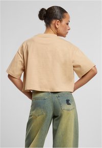 Lühikeste varrukatega pehme puuvillane beež T-särk, mida kantakse sinise-rohelise gradientvärvi teksadega, millel on iseloomulik taskulogo.