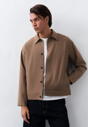 Muž v hnedej bunde na gombíky cez bielu košeľu a tmavé džínsy, stojaci pred svetlosivým pozadím, pozerajúci sa do strany.