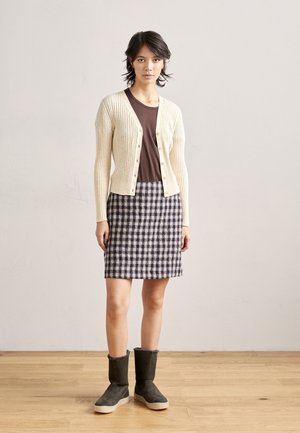 Femme portant un cardigan côtelé crème sur une chemise marron avec une jupe à carreaux marine et crème mi-longue jusqu'aux genoux et des bottes mi-mollet gris foncé.