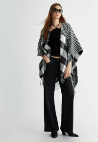 Poncho bianco e nero con motivo a quadri e bordi frangiati, indossato sopra un top aderente nero e abbinato a jeans a gamba larga neri e tacchi a punta.