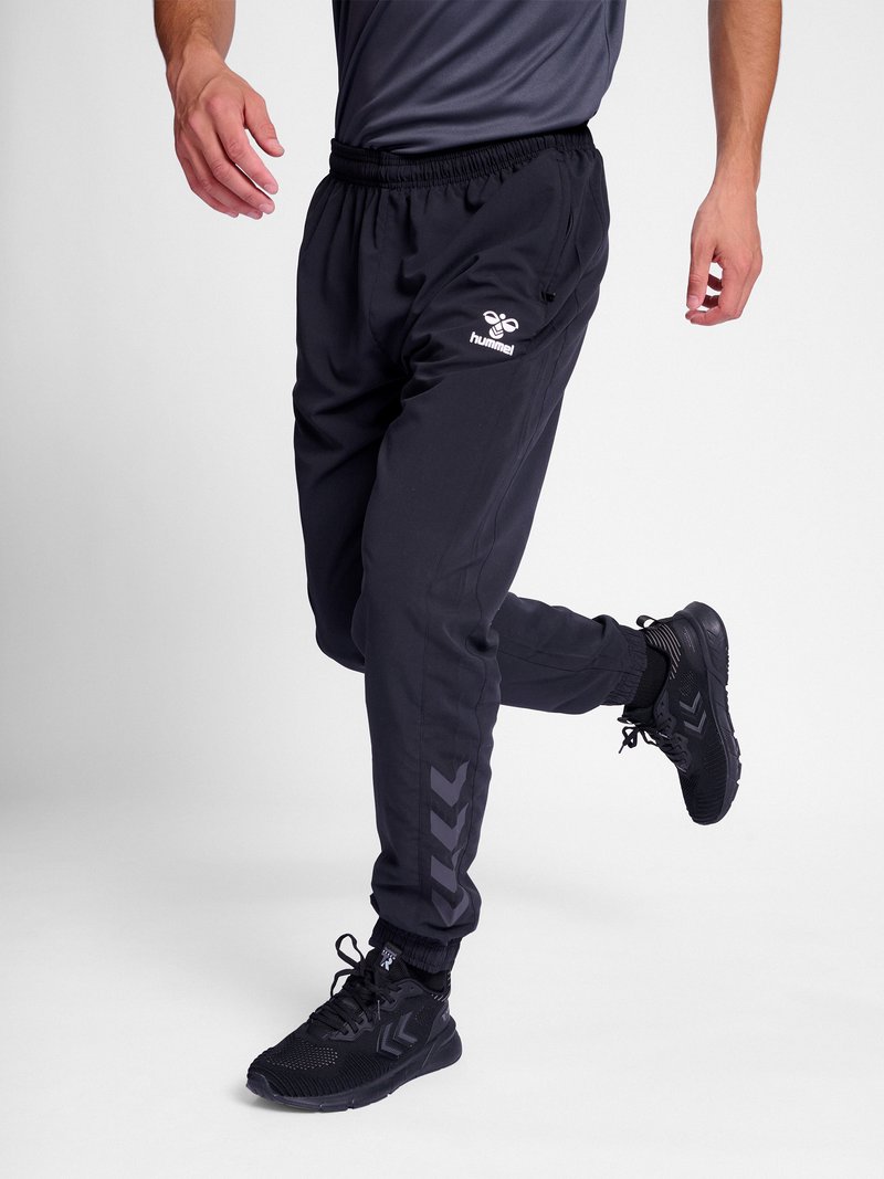 Person trägt schwarze Hummel-Jogginghosen und schwarze Sportsneaker, gezeigt von der Taille bis zu den Füßen in einer Gehbewegung.